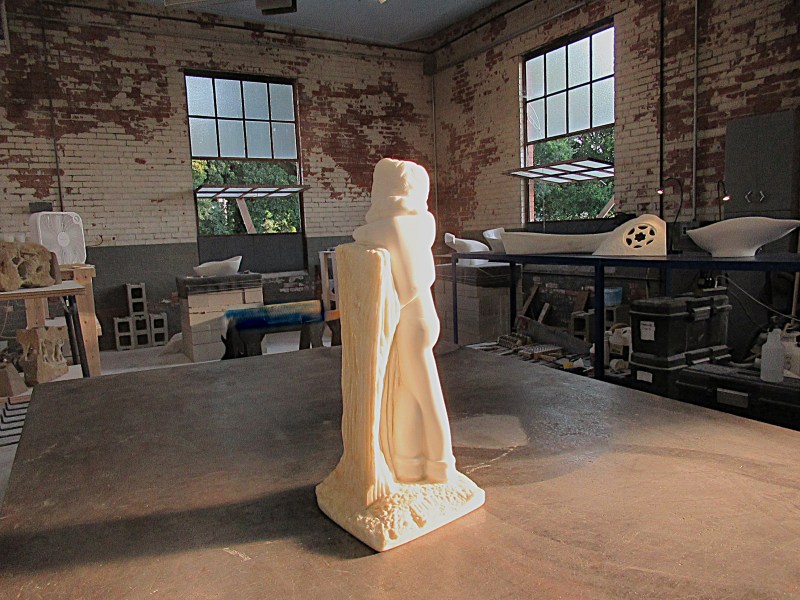 A white sculpture of a figure standing by a log on a wooden table, set in an art studio with brick walls and large windows.