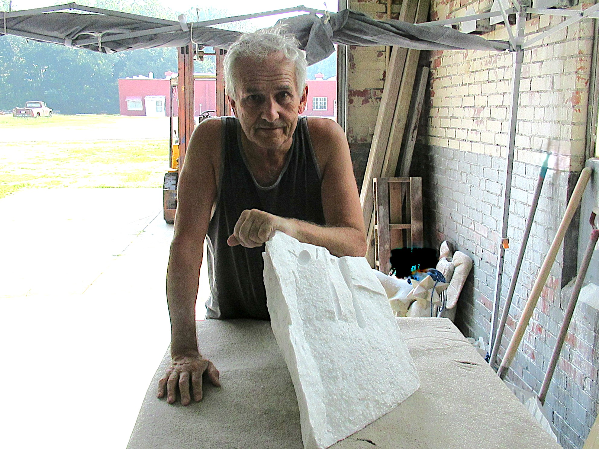 A middle-aged man with gray hair is leaning on a stone sculpture in a workshop setting, with various tools and materials in the background.